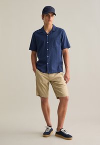 Chemise boutonnée à manches courtes navy en tissu texturé, shorts kaki et baskets navy avec lacets blancs ; portant une casquette navy assortie.