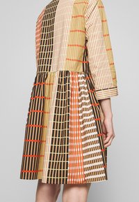 Gingham-Kleid mit einer Mischung aus Farben: beigen, braunen, orangefarbenen und grünen Streifen. Es hat lange Ärmel und einen gerafften Rock.