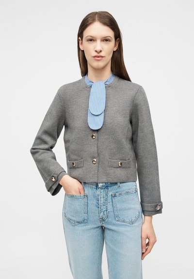 Γυναίκα που φορά γκρι πλεκτή ζακέτα με χρυσά κουμπιά, μπλε γραμμωτή γραβάτα και ανοιχτόχρωμο μπλε τζιν παντελόνι με εμπρόσθιες τσέπες, στάση με το χέρι στην τσέπη.