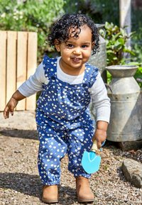 Blaue Blumen-Overalls mit Rüscheträgern, kombiniert mit einem weißen Langarmshirt und braunen Stiefeln. Hält eine hellblaue Plastikschelle.