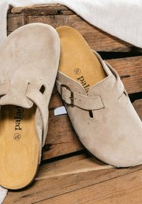 Een paar beige suède instapsloffen met gespen en kleine gouden studs, geplaatst op een houten oppervlak met een stuk stof ernaast.