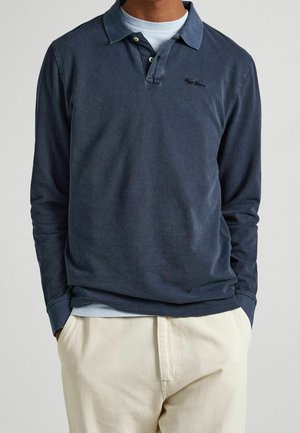 Mężczyzna ubrany w granatową, długorękawową koszulkę polo na jasnoniebieskim t-shircie oraz beżowe spodnie, ze włożonymi rękami do kieszeni, na neutralnym tle.