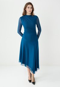 Robe turquoise avec de longues manches en maille, corsage ajusté et jupe asymétrique fluide. Tissu lisse avec un ourlet transparent. Escarpins noirs à talons pointus.