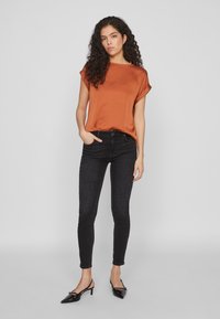 Top in satin color ruggine con maniche corte arrotolate, abbinato a jeans skinny neri e décolleté neri con tacco. Sfondo semplice.