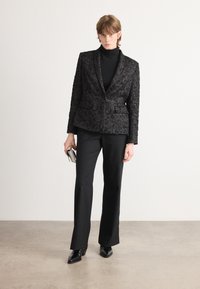 Zwarte geprinte blazer met een textuur, gecombineerd met een zwarte coltrui, strakke zwarte broek en elegante zwarte hakken.