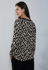 Černá blůza s abstraktním krémovým zigzag vzorem, dlouhými rukávy, uvolněným střihem a kulatým výstřihem. Hladká látka s mírným leskem.