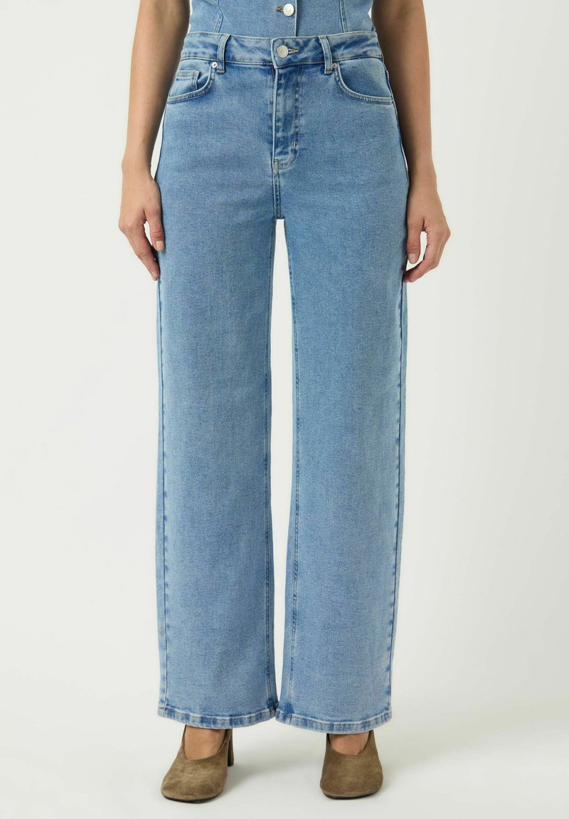 Nainen, joka pukeutuu vaaleansinisiin leveälahkeisiin farkkuihin ja ruskeisiin korkeakorkoisiin kenkiin, seisoo yksinkertaisen vaalean taustan edessä.