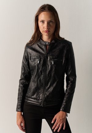 Jeune femme aux longs cheveux bruns portant une veste en cuir noire zippée et un pantalon noir, debout devant un fond clair uni.
