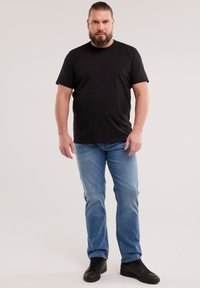 Habemega mees, kellel on seljas must T-särk, sinised teksad ja mustad tossud, seisab ükslasel taustal.