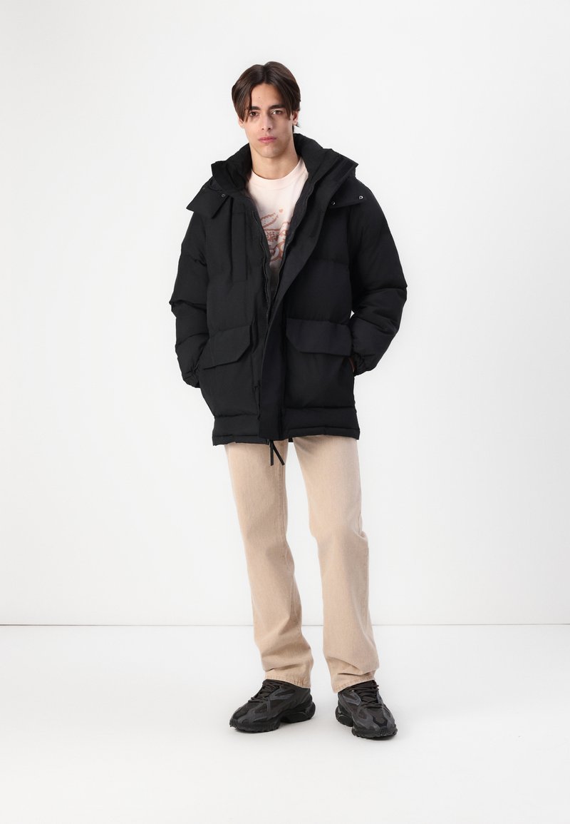 Veste noire matelassée avec capuche, deux poches avant et cordon de serrage à l'ourlet, portée sur un t-shirt clair et un pantalon beige.
