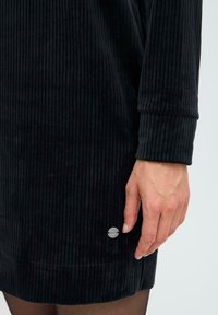 Zwarte geribbelde corduroy jurk met een relaxed fit, lange mouwen en een klein metalen logo detail nabij de zoom. De textuur lijkt zacht.