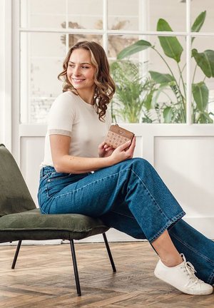 Model wearing a cream short-sleeve shirt and blue high-waisted jeans, holding a brown patterned wallet, seated on a green chair.