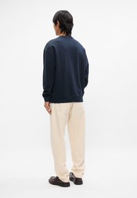 Ναυτικό βαμβακερό φούτερ με στρογγυλή λαιμόκοψη και ραμμένα μανίκια, φορεμένο με μπεζ παντελόνι και μαύρες λοφέρες, εικονιζόμενο από πίσω.