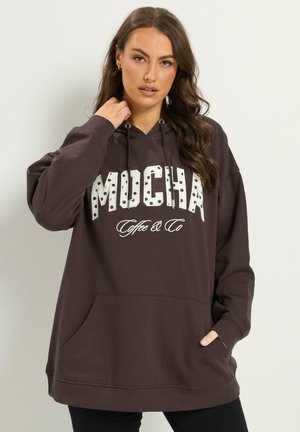 Femme aux longs cheveux bruns portant un sweat à capuche marron foncé avec le texte "MOCHA Coffee & Co", une main dans la poche, l’autre près du cou.