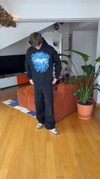 Fekete kapucnis pulóver kék grafikával, laza fekete nadrággal és ezüst cipővel párosítva. A háttérben beltéri növények és bútorok láthatók.