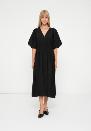 Frau in einem schwarzen Midikleid mit puffärmel und V-Ausschnitt, kombiniert mit schwarzen, spitzen Slingback-High Heels, vor einem weißen Hintergrund stehend.