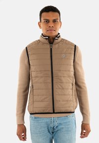 Gilet rembourré beige avec un col montant, texture matelassée, fermeture éclair à l'avant et bordure bleu marine. Assorti à un pull beige et un jean en denim clair.