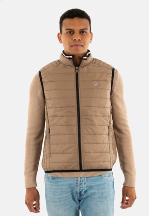 Gilet rembourré beige avec un col montant, texture matelassée, fermeture éclair à l'avant et bordure bleu marine. Assorti à un pull beige et un jean en denim clair.