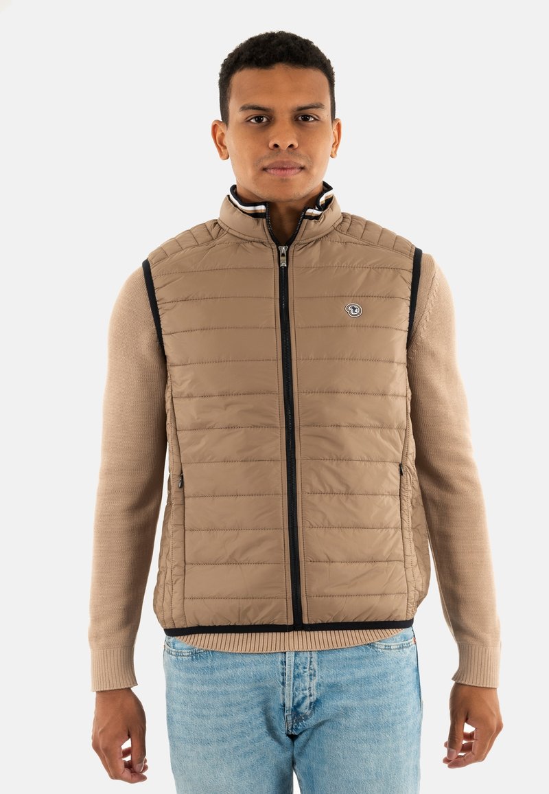 Gilet rembourré beige avec un col montant, texture matelassée, fermeture éclair à l'avant et bordure bleu marine. Assorti à un pull beige et un jean en denim clair.