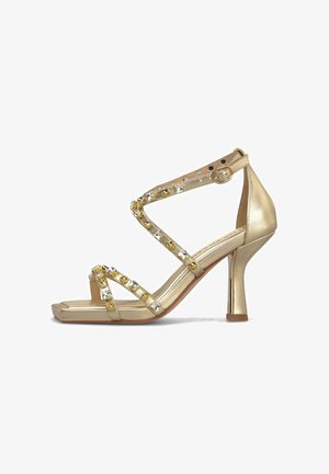 Sandale à talon haut dorée avec bout carré, brides croisées ornées de clous dorés et transparents, et fermeture à boucle à la cheville.