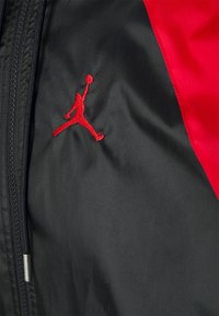 Černá a červená bunda s hladkou texturou, která má vyšívané červené logo Jumpman a zapínání na zip na levé straně.