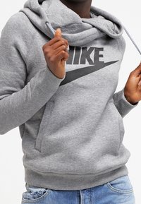 Grå sweatshirt med huva, med en stor svart Nike-logotyp på framsidan, kengurufickor och ribbade ärmslut och fåll.