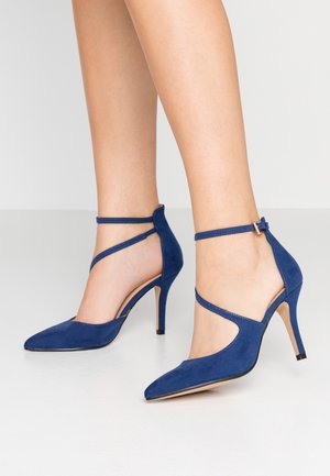 Mørkeblå ruskinds højhælede sko med spids tå og ankelstropper. Har et åbent sideskær og en slank 10 cm stilethæl.