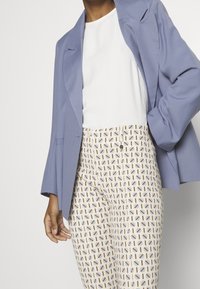 Lyseblå blazer over en hvid skjorte, parret med mønstrede beige bukser med geometriske former i blå og brune nuancer.