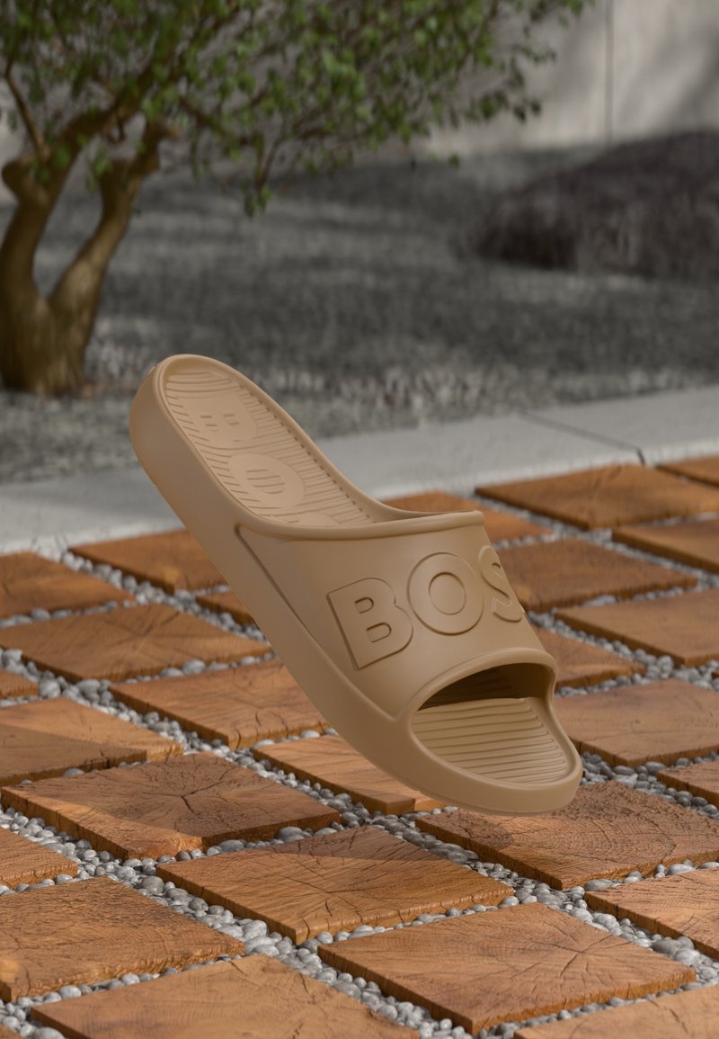 Semelle en caoutchouc beige avec une large sangle portant le mot "BOSS" en relief. Semelle intérieure rainurée. Flottant au-dessus de carrelages bruns texturés avec des galets gris.