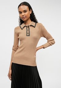 Ριγέ μπεζ πουλόβερ με μαύρη επένδυση στον γιακά και κουμπί, σε συνδυασμό με μια μαύρη πλισέ φούστα. Η υφή είναι λεία και εφαρμοστή.