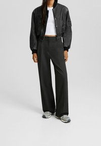 Svart kort bomberjacka med ribbade manschetter, kombinerad med högmidjade mörkgrå vida byxor och grå sneakers med svarta detaljer.