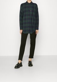 Karierte Knopfleiste-Hemd in Dunkelgrün und Marineblau, mit einer Brusttasche und langen Ärmeln. Kombiniert mit schwarzen, taillierten Hosen und glänzenden Slippern.