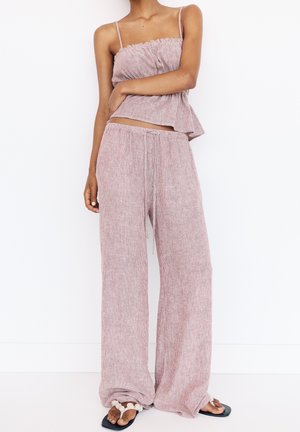 Set de îmbrăcăminte de relaxare ușor, cu model ribbed, de un roz moale, cu un top scurt cu volane și pantaloni largi cu șnur. Sandale cu accente florale.