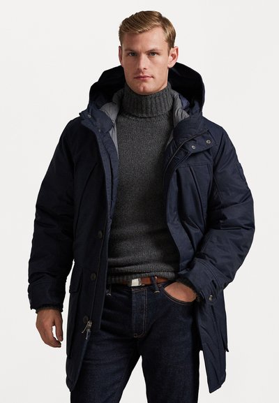 Parka blu navy con cappuccio removibile, chiusura con zip e bottoni, e fodera grigia. Indossata sopra un maglione scuro a trama, abbinata a jeans scuri.