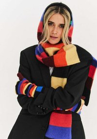 Wielokolorowy, prążkowany szal wełniany noszony z czarną marynarką, charakteryzujący się odważnymi kolorami takimi jak czerwony, niebieski, żółty i pomarańczowy oraz fakturą prążkowaną.