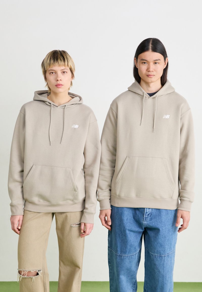 Felpe con cappuccio beige con tasche frontali, indossate da due persone. Le felpe presentano un piccolo logo bianco. Una indossa pantaloni beige, l'altra in denim.