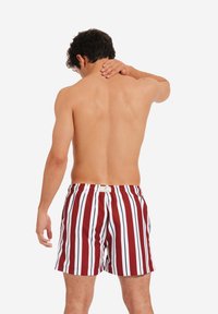 Costumi da bagno a righe rosse e bianche con vita elastica e una piccola etichetta; il modello è mostrato di schiena, con la pelle nuda visibile.