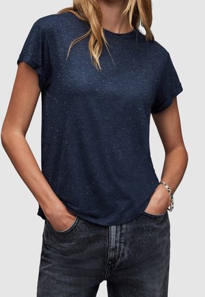 Kobieta w granatowym, cętkowanym, krótkim rękawem t-shircie wpuszczonym w czarne, prane jeansy, z rękami w kieszeniach i srebrną bransoletką na lewym nadgarstku.
