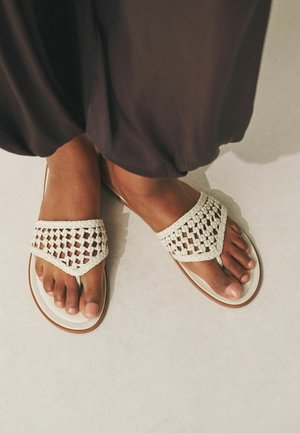 Lábak bézs szandálban, fonott pántokkal, világos padlón állnak, részben sötét bő nadrággal fedve.