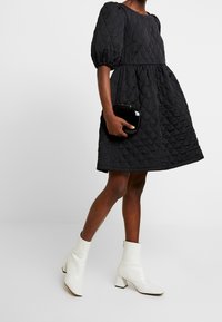 Schwarzes gesteppte Kleid mit kurzen, gepufften Ärmeln und einem ausgestellten Rock, kombiniert mit einer schwarzen Clutch und weißen, klobigen, geschnürten Ankle Boots.