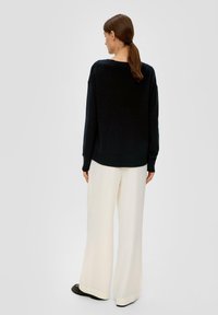 Schwarzer Strickpullover mit überschnittenen Schultern und Rundhalsausschnitt, kombiniert mit weiten cremefarbenen Hosen. Glatte Textur, tailliert an der Taille, insgesamt lockere Passform.
