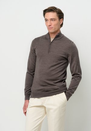Donkerbruine wollen pullover trui met een half-rits kraag, lange mouwen en een strakke pasvorm. Gepaard met crèmekleurige broeken.