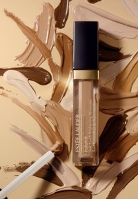 Estee Lauder Futurist Soft Touch Brightening Skincare in een glazen tube met een zwarte dop, geplaatst op draaiende beige en bruine crèmeswatches.