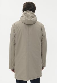 Vyras, vilkintis smėlio spalvos žiemos striukę su užtrauktuku, matomas iš nugaros prieš baltą foną, rankos šonuose.