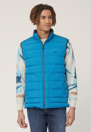 Gilet trapuntato blu con colletto alto, chiusura frontale con zip e tasche laterali. Indossato sopra una maglietta a maniche lunghe con fantasia.