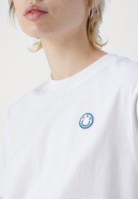 Hvid bomulds t-shirt med et broderet blåt smiley ansigtspatch med bogstaverne "H," "G," og "O" nær halsudskæringen.