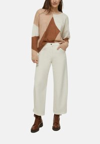 Maglione lavorato a maglia in beige, marrone e crema con motivo geometrico; vestibilità ampia. Pantaloni bianchi a gamba larga; stivali marroni con lacci e suole spesse.