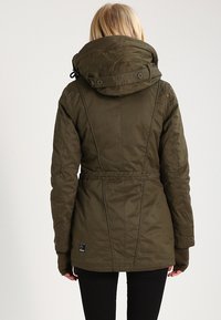 Žena s blond vlasy, oblečená v olivově zelené zimní bundě s kapucí, černých kalhotách a hnědých rukavicích, viděná zezadu.