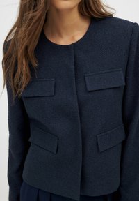 Tamsiai mėlyna tekstūruota striukė su apvaliu iškirpimu, dviem priekinėmis kišenėmis ir ilgom rankovėmis, pasižyminti švariu, pritaikytu dizainu.