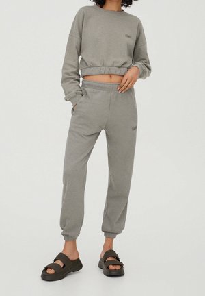 Persoon draagt lichtgrijs cropped sweatshirt en bijpassende joggerbroek met zwarte slip-on sandalen tegen een effen achtergrond.